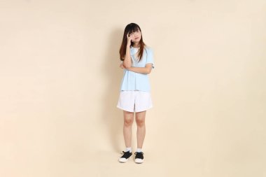 Açık mavi polo tişört giyen, bej arka planda beyaz şortlu genç bir Asyalı kadının vücut portresi. Üniversite öğrencisi, Gen Z