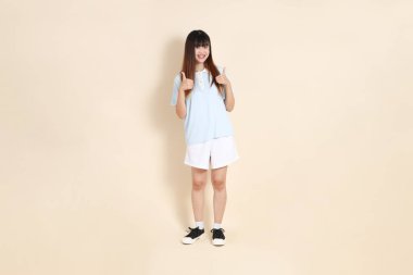 Açık mavi polo tişört giyen genç bir Asyalı kadının tüm vücut portresi ve beyaz şort, baş parmaklar yukarı, tamam ya da bej bir arka planda onaylayın. Üniversite öğrencisi, Gen Z