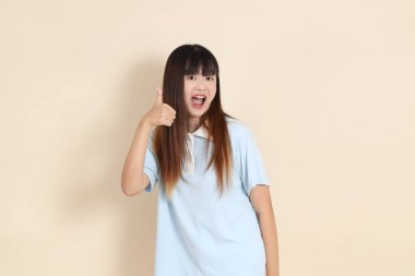 Orta boy Asyalı bir kadının açık mavi polo tişörtü ve beyaz şort giydiği bir fotoğraf. Başparmaklar yukarı, tamam mı? Ya da bej bir arka plan. Üniversite öğrencisi, Gen Z