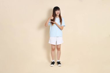 Açık mavi polo tişört giyen genç bir Asyalı kadının tüm vücut portresi ve beyaz şort, baş parmaklar yukarı, tamam ya da bej bir arka planda onaylayın. Üniversite öğrencisi, Gen Z