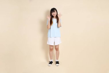 Açık mavi polo tişört giyen genç bir Asyalı kadının tüm vücut portresi ve beyaz şort, baş parmaklar yukarı, tamam ya da bej bir arka planda onaylayın. Üniversite öğrencisi, Gen Z