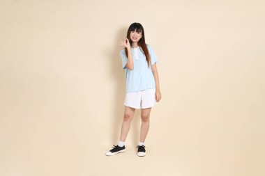 Açık mavi polo tişört giyen genç bir Asyalı kadının tüm vücut portresi ve beyaz şort, baş parmaklar yukarı, tamam ya da bej bir arka planda onaylayın. Üniversite öğrencisi, Gen Z