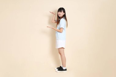 Açık mavi polo tişört giyen genç bir Asyalı kadının tam vücut portresi ve beyaz şort, Ölçü Sembolü, küçük, büyük, düz bir bej arka planda. Üniversite öğrencisi, Gen Z