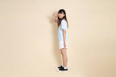 Açık mavi polo tişört giyen genç bir Asyalı kadının tam vücut portresi ve beyaz şort, Ölçü Sembolü, küçük, büyük, düz bir bej arka planda. Üniversite öğrencisi, Gen Z