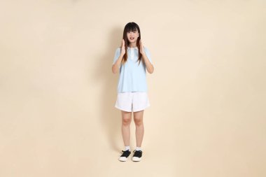 Açık mavi polo tişört giyen genç bir Asyalı kadının tam vücut portresi ve beyaz şort, Ölçü Sembolü, küçük, büyük, düz bir bej arka planda. Üniversite öğrencisi, Gen Z