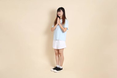 Açık mavi polo tişört giyen, beyaz şortlu, bej arka planda duran genç bir Asyalı kadının vücut portresi. Üniversite öğrencisi, Gen Z