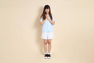 Açık mavi polo tişört giyen genç bir Asyalı kadının vücut portresi ve beyaz şort, bej arka planda dua ve huzur. Üniversite öğrencisi, Gen Z