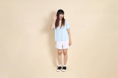 Açık mavi polo tişörtlü, beyaz şortlu genç bir Asyalı kadın bej arka planda karşılanıyor. Üniversite öğrencisi, Gen Z, Summer