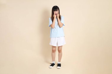 Açık mavi polo tişörtlü genç bir Asyalı kadın ve beyaz şortlu, hayal kırıklığına uğramış, bej bir arka planda ağlayan. Üniversite öğrencisi, Gen Z, Summer