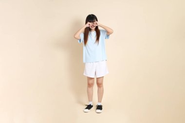 Açık mavi polo tişörtlü genç bir Asyalı kadın ve beyaz şortlu, hayal kırıklığına uğramış, bej bir arka planda ağlayan. Üniversite öğrencisi, Gen Z, Summer