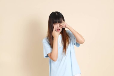 Açık mavi polo tişörtlü genç bir Asyalı kadın ve beyaz şortlu, hayal kırıklığına uğramış, bej bir arka planda ağlayan. Üniversite öğrencisi, Gen Z, Summer