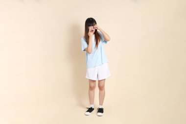 Açık mavi polo tişörtlü genç bir Asyalı kadın ve beyaz şortlu, hayal kırıklığına uğramış, bej bir arka planda ağlayan. Üniversite öğrencisi, Gen Z, Summer