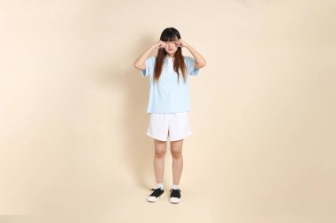 Açık mavi polo tişörtlü genç bir Asyalı kadın ve beyaz şortlu, hayal kırıklığına uğramış, bej bir arka planda ağlayan. Üniversite öğrencisi, Gen Z, Summer