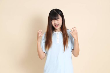 Açık mavi polo tişörtlü genç bir Asyalı kadın ve beyaz şortlu, kızgın ve bej arka planda çığlık atan bir kadın. Üniversite öğrencisi, Gen Z, Summer