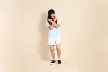 Açık mavi polo tişörtlü, beyaz şortlu, bej arka plandan korkan genç bir Asyalı kadın. Üniversite öğrencisi, Gen Z, Summer