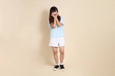 Açık mavi polo tişörtlü, beyaz şortlu, bej arka plandan korkan genç bir Asyalı kadın. Üniversite öğrencisi, Gen Z, Summer