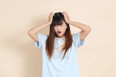 Açık mavi polo tişörtlü genç bir Asyalı kadın, ve beyaz şortlu, baş ağrısı ya da stresi olan düz bir bej arka plan. Üniversite öğrencisi, Gen Z, Summer