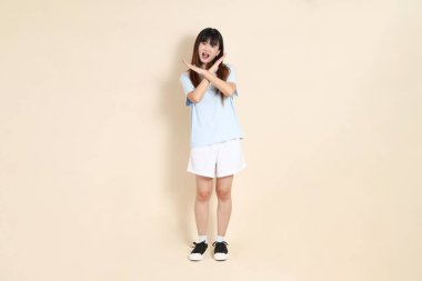 Açık mavi polo tişörtlü, beyaz şortlu genç bir Asyalı kadın, dur, reddediyor, düz bir bej arka plana hayır diyor. Üniversite öğrencisi, Gen Z, Summer