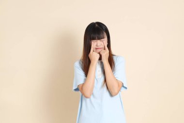 Açık mavi polo tişörtlü genç bir Asyalı kadın ve beyaz şortlu, hayal kırıklığına uğramış, bej bir arka planda ağlayan. Üniversite öğrencisi, Gen Z, Summer