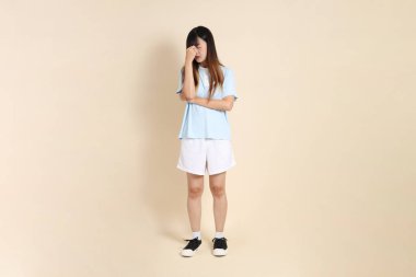 Açık mavi polo tişörtlü genç bir Asyalı kadın, ve beyaz şortlu, baş ağrısı ya da stresi olan düz bir bej arka plan. Üniversite öğrencisi, Gen Z, Summer