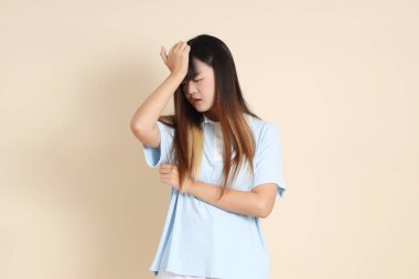 Açık mavi polo tişörtlü genç bir Asyalı kadın, ve beyaz şortlu, baş ağrısı ya da stresi olan düz bir bej arka plan. Üniversite öğrencisi, Gen Z, Summer