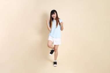 Açık mavi polo tişört giyen genç Asyalı kadın ve beyaz şort, evet, heyecanlı, bej arka planda kutlama yapıyor. Üniversite öğrencisi, Gen Z, Summer