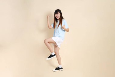 Açık mavi polo tişört giyen genç Asyalı kadın ve beyaz şort, evet, heyecanlı, bej arka planda kutlama yapıyor. Üniversite öğrencisi, Gen Z, Summer