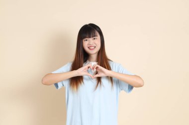 Açık mavi polo tişörtlü genç bir Asyalı kadın, ve beyaz şort, kalp şeklinde, mini kalp, düz bir bej arka planda aşk. Üniversite öğrencisi, Gen Z, Summer