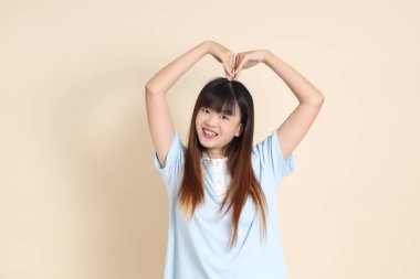 Açık mavi polo tişörtlü genç bir Asyalı kadın, ve beyaz şort, kalp şeklinde, mini kalp, düz bir bej arka planda aşk. Üniversite öğrencisi, Gen Z, Summer