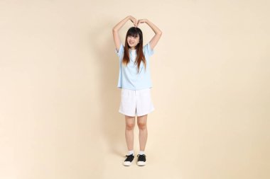 Açık mavi polo tişörtlü genç bir Asyalı kadın, ve beyaz şort, kalp şeklinde, mini kalp, düz bir bej arka planda aşk. Üniversite öğrencisi, Gen Z, Summer