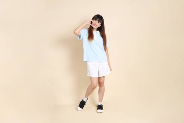 Açık mavi polo tişörtlü genç bir Asyalı kadın, ve beyaz şort, kalp şeklinde, mini kalp, düz bir bej arka planda aşk. Üniversite öğrencisi, Gen Z, Summer