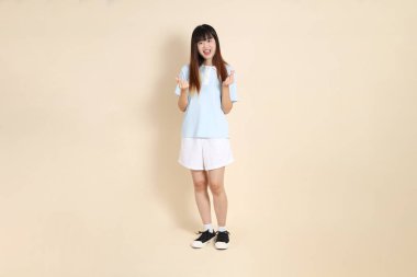 Açık mavi polo tişörtlü genç bir Asyalı kadın, ve beyaz şort, kalp şeklinde, mini kalp, düz bir bej arka planda aşk. Üniversite öğrencisi, Gen Z, Summer