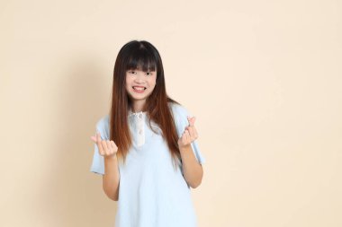 Açık mavi polo tişörtlü genç bir Asyalı kadın, ve beyaz şort, kalp şeklinde, mini kalp, düz bir bej arka planda aşk. Üniversite öğrencisi, Gen Z, Summer