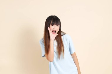 Açık mavi polo tişörtlü, beyaz şortlu, bağıran, bej arka planda megafon tutan genç bir Asyalı kadın. Üniversite öğrencisi, Gen Z, Summer
