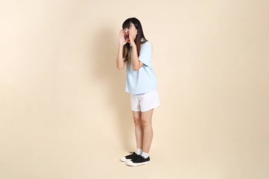 Açık mavi polo tişörtlü, beyaz şortlu, bağıran, bej arka planda megafon tutan genç bir Asyalı kadın. Üniversite öğrencisi, Gen Z, Summer
