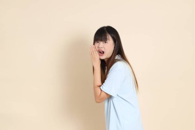 Açık mavi polo tişörtlü, beyaz şortlu, bağıran, bej arka planda megafon tutan genç bir Asyalı kadın. Üniversite öğrencisi, Gen Z, Summer