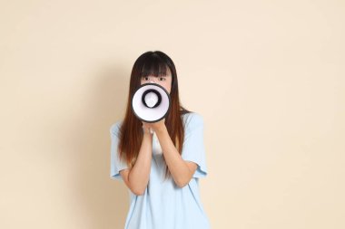 Açık mavi polo tişörtlü, beyaz şortlu, bağıran, bej arka planda megafon tutan genç bir Asyalı kadın. Üniversite öğrencisi, Gen Z, Summer