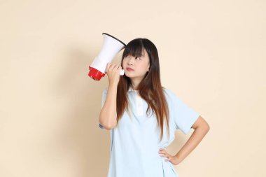 Açık mavi polo tişörtlü, beyaz şortlu, bağıran, bej arka planda megafon tutan genç bir Asyalı kadın. Üniversite öğrencisi, Gen Z, Summer