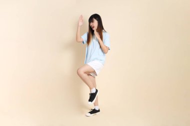 Açık mavi polo tişörtlü, beyaz şortlu genç bir Asyalı kadın bej bir arka plana karşı savaşıyor. Üniversite öğrencisi, Gen Z, Summer