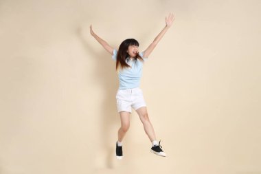 Açık mavi polo tişörtlü, beyaz şortlu, bej arka planda zıplayan genç Asyalı bir kadın. Üniversite öğrencisi, Gen Z, Summer