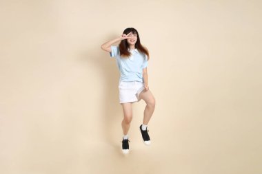Açık mavi polo tişörtlü, beyaz şortlu, bej arka planda zıplayan genç Asyalı bir kadın. Üniversite öğrencisi, Gen Z, Summer