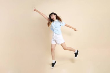 Açık mavi polo tişörtlü, beyaz şortlu, bej arka planda zıplayan genç Asyalı bir kadın. Üniversite öğrencisi, Gen Z, Summer