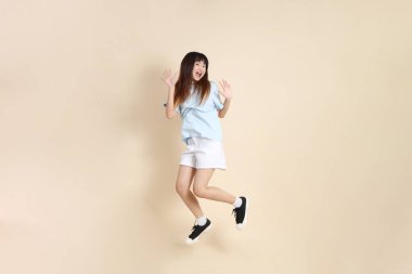 Açık mavi polo tişörtlü, beyaz şortlu, bej arka planda zıplayan genç Asyalı bir kadın. Üniversite öğrencisi, Gen Z, Summer