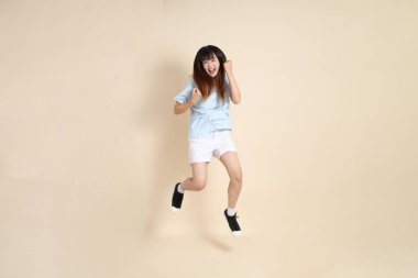 Açık mavi polo tişörtlü, beyaz şortlu, bej arka planda zıplayan genç Asyalı bir kadın. Üniversite öğrencisi, Gen Z, Summer