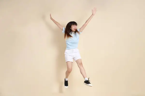Açık mavi polo tişörtlü, beyaz şortlu, bej arka planda zıplayan genç Asyalı bir kadın. Üniversite öğrencisi, Gen Z, Summer