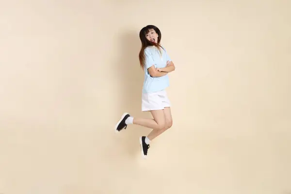 Açık mavi polo tişörtlü, beyaz şortlu, bej arka planda zıplayan genç Asyalı bir kadın. Üniversite öğrencisi, Gen Z, Summer