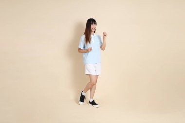 Açık mavi polo tişörtlü, beyaz şortlu, bej arka planda yürüyen genç Asyalı bir kadın. Üniversite öğrencisi, Gen Z, Summer