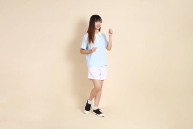 Açık mavi polo tişörtlü, beyaz şortlu, bej arka planda yürüyen genç Asyalı bir kadın. Üniversite öğrencisi, Gen Z, Summer
