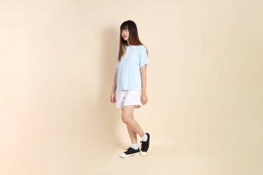 Açık mavi polo tişörtlü, beyaz şortlu, bej arka planda yürüyen genç Asyalı bir kadın. Üniversite öğrencisi, Gen Z, Summer