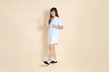 Açık mavi polo tişörtlü, beyaz şortlu, bej arka planda yürüyen genç Asyalı bir kadın. Üniversite öğrencisi, Gen Z, Summer